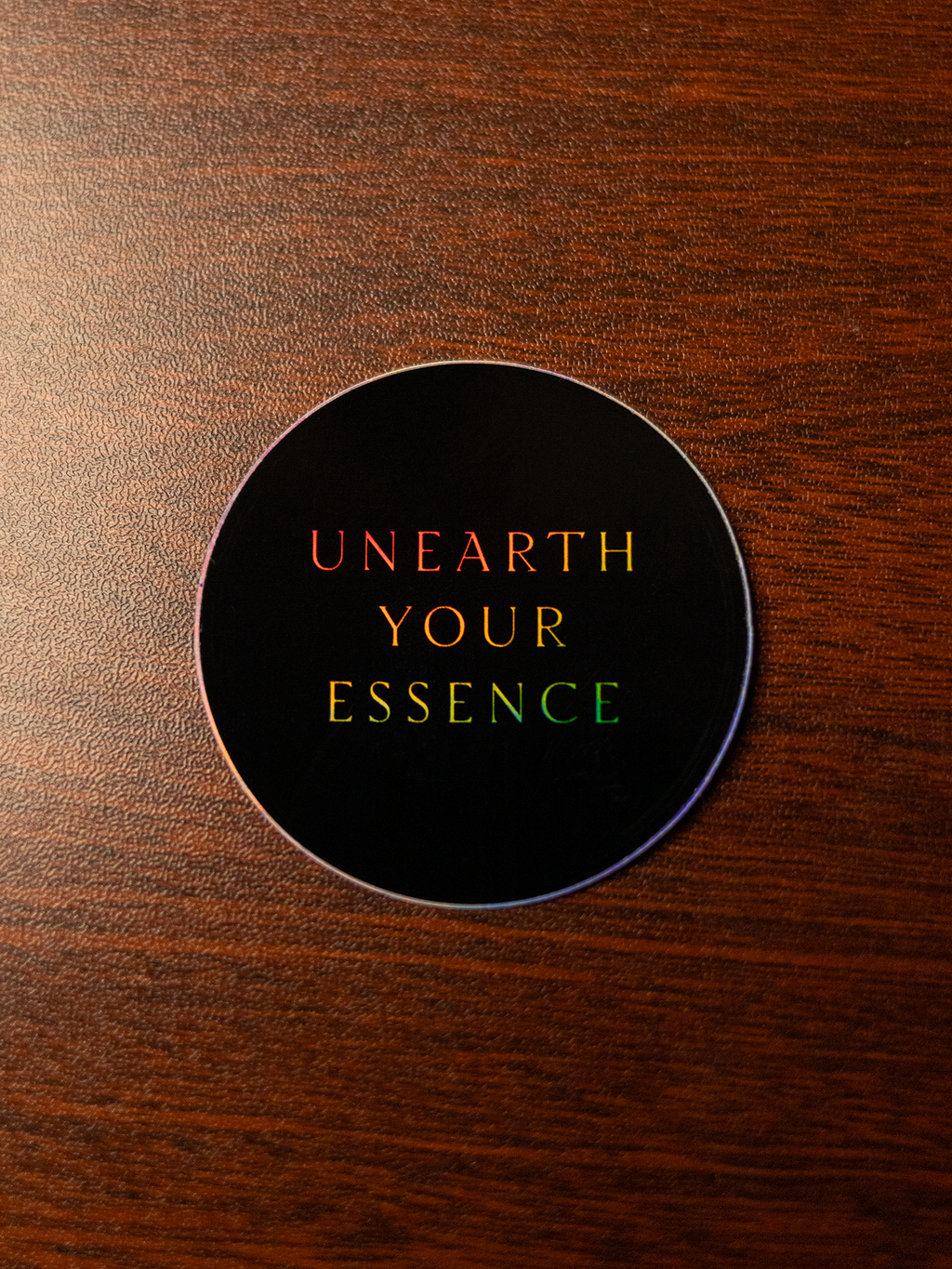 Round black sticker with the words "Unearth Your Essence" sitting on a wooden surface