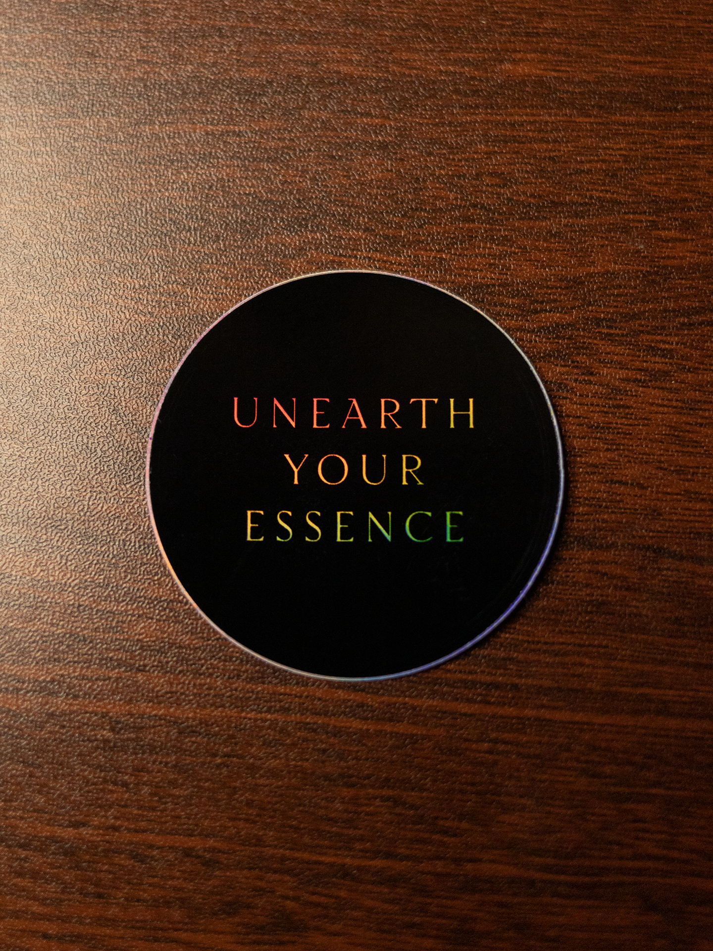Round black sticker with the words "Unearth Your Essence" sitting on a wooden surface